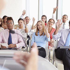 Office environment with young adults who are sitting at their desks facing the professor raising their hands with questions.